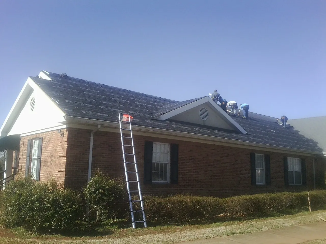 Crew tearing shingles off a brick ranch roof with a ladder set — Emergency Roof Repair work by our crew in Kinston
